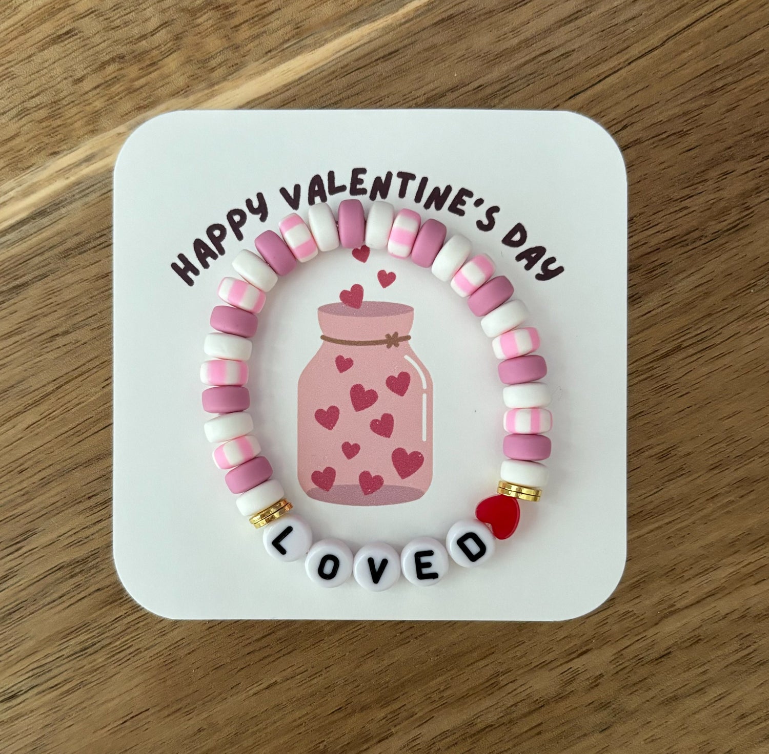 A multicolored beaded bracelet with 'HAPPY Valentine's DAY' written on it, featuring pink and red jade beads, gold plating, and acrylic letter beads on an elastic cord.