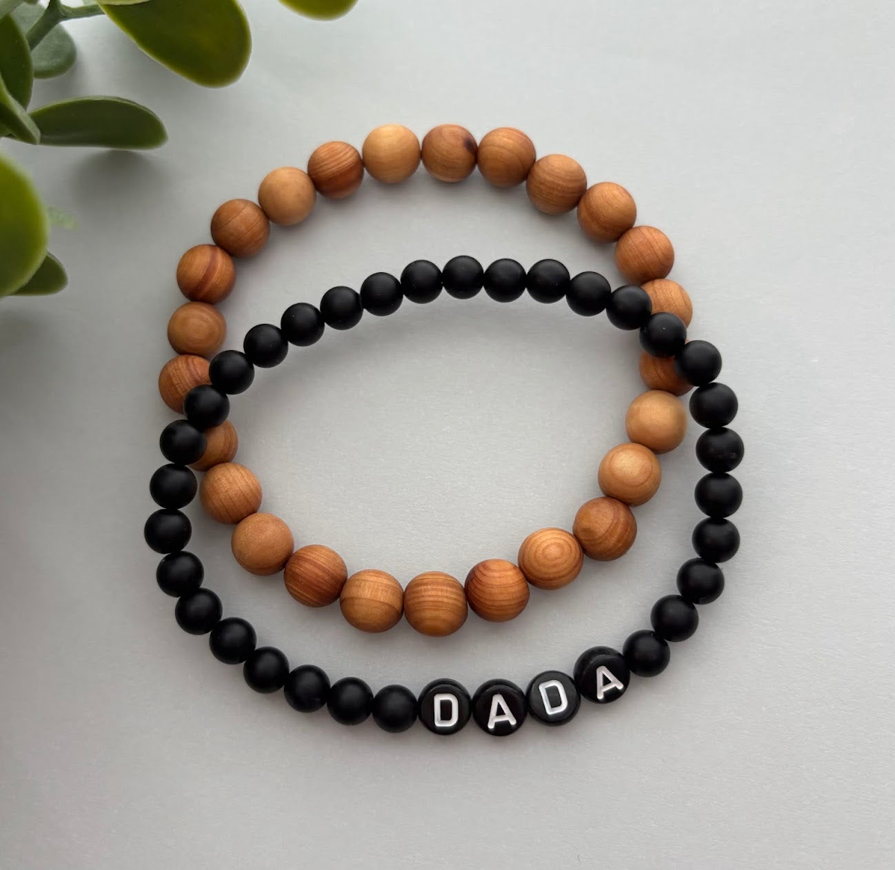 Two bracelets, one with wooden beads and the other with black beads and 'DADA' letters, on a light surface.
