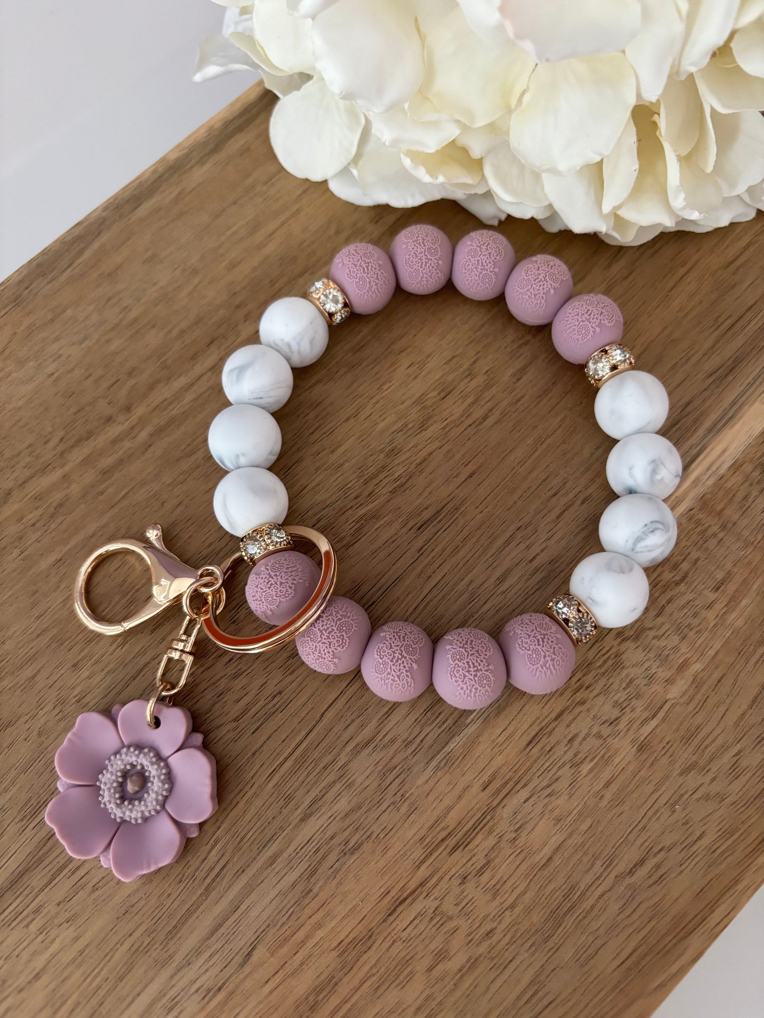 A mauve and white marble keychain wristlet with a gold-toned clasp and a mauve poppy bead, displayed on a wooden surface next to a white flower.