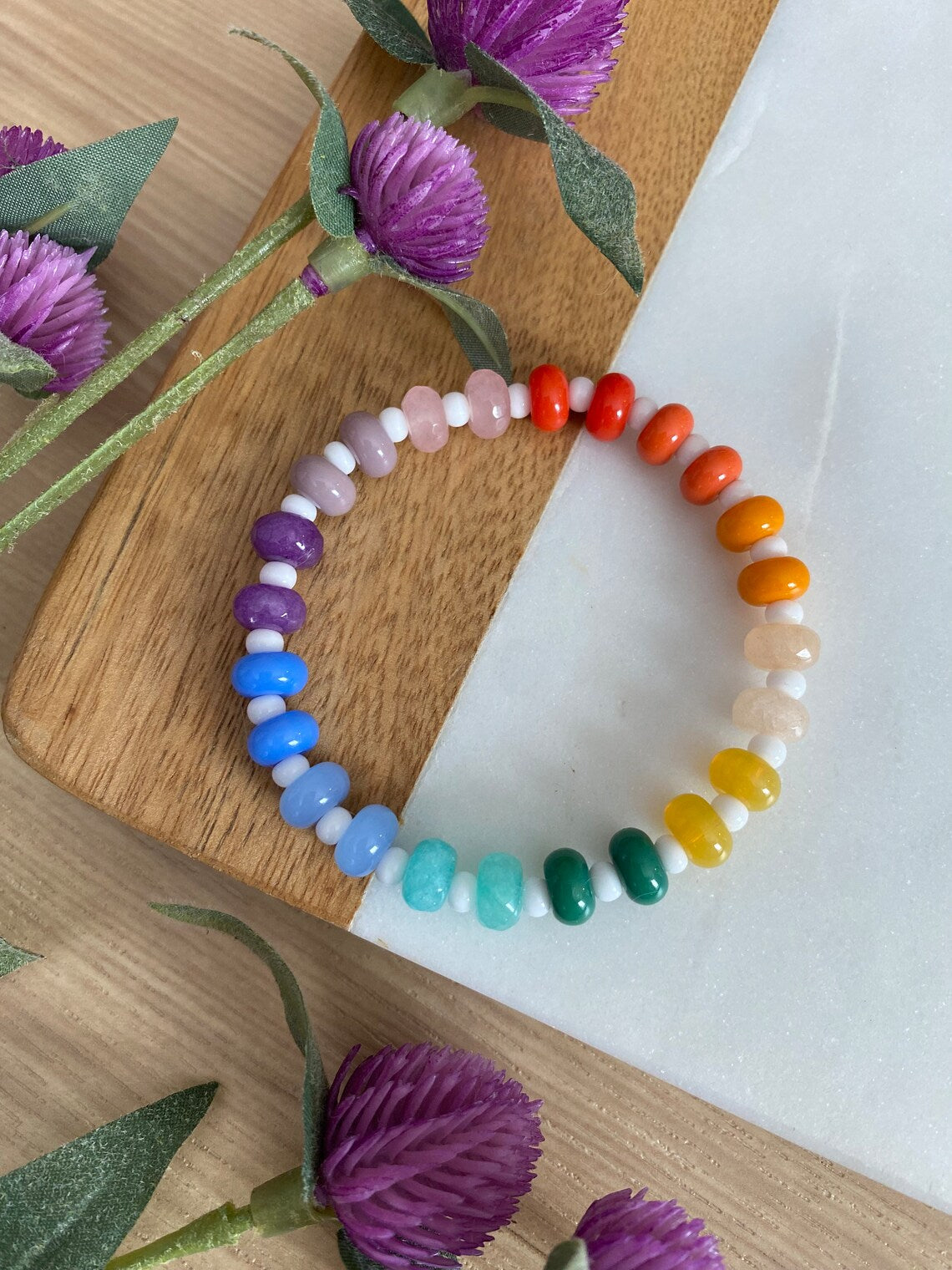 A multicolored beaded bracelet with Jade and glass beads, displayed on a wooden surface with purple flowers and a sprig of greenery in the background.