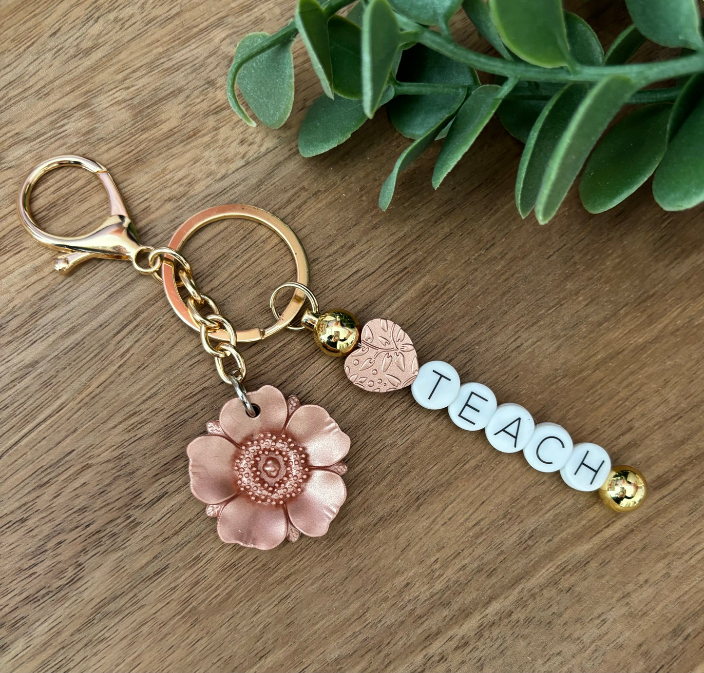 A keychain with a floral design and the word 'Teach' written on it, displayed on a wooden surface.