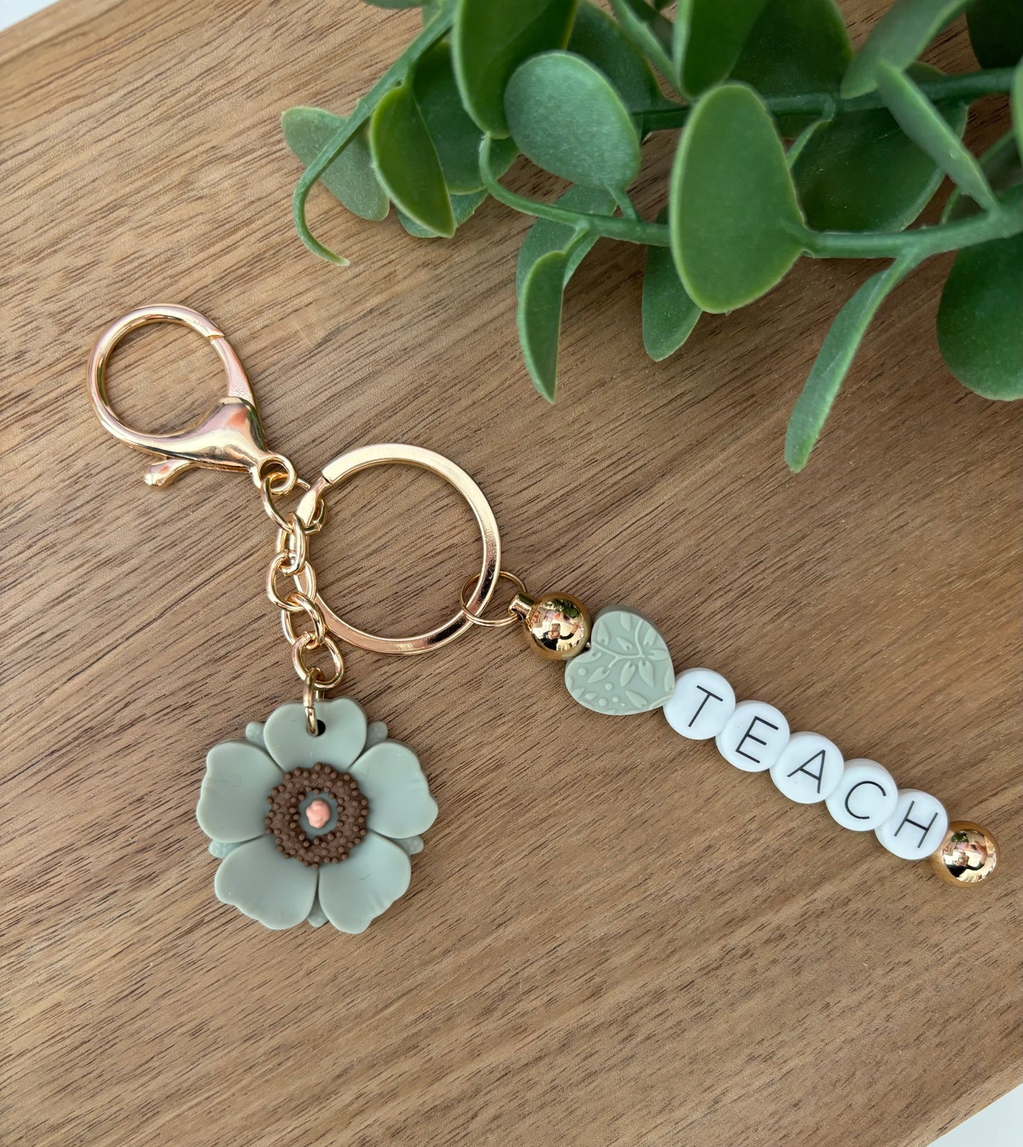 A keychain with a floral design and the word 'Teach' written on it, displayed on a wooden surface.