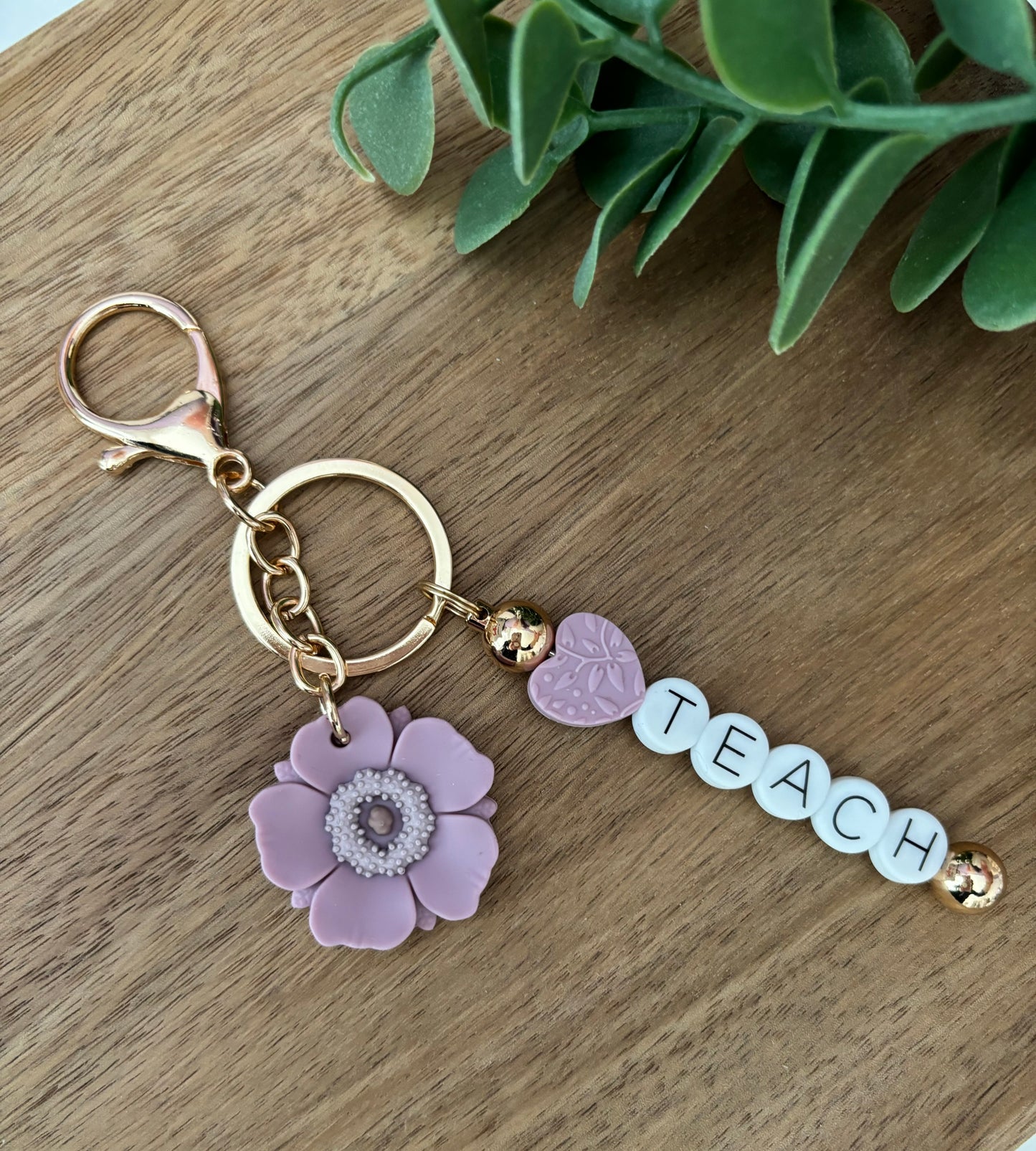 A keychain with a floral design and the word 'Teach' written on it, displayed on a wooden surface.