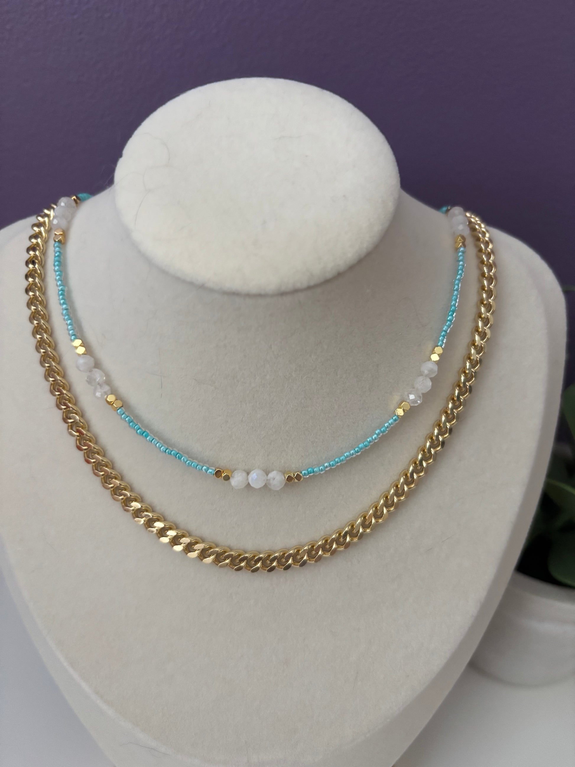 Gold chain bracelet with blue and white beads on a white stand against a purple background