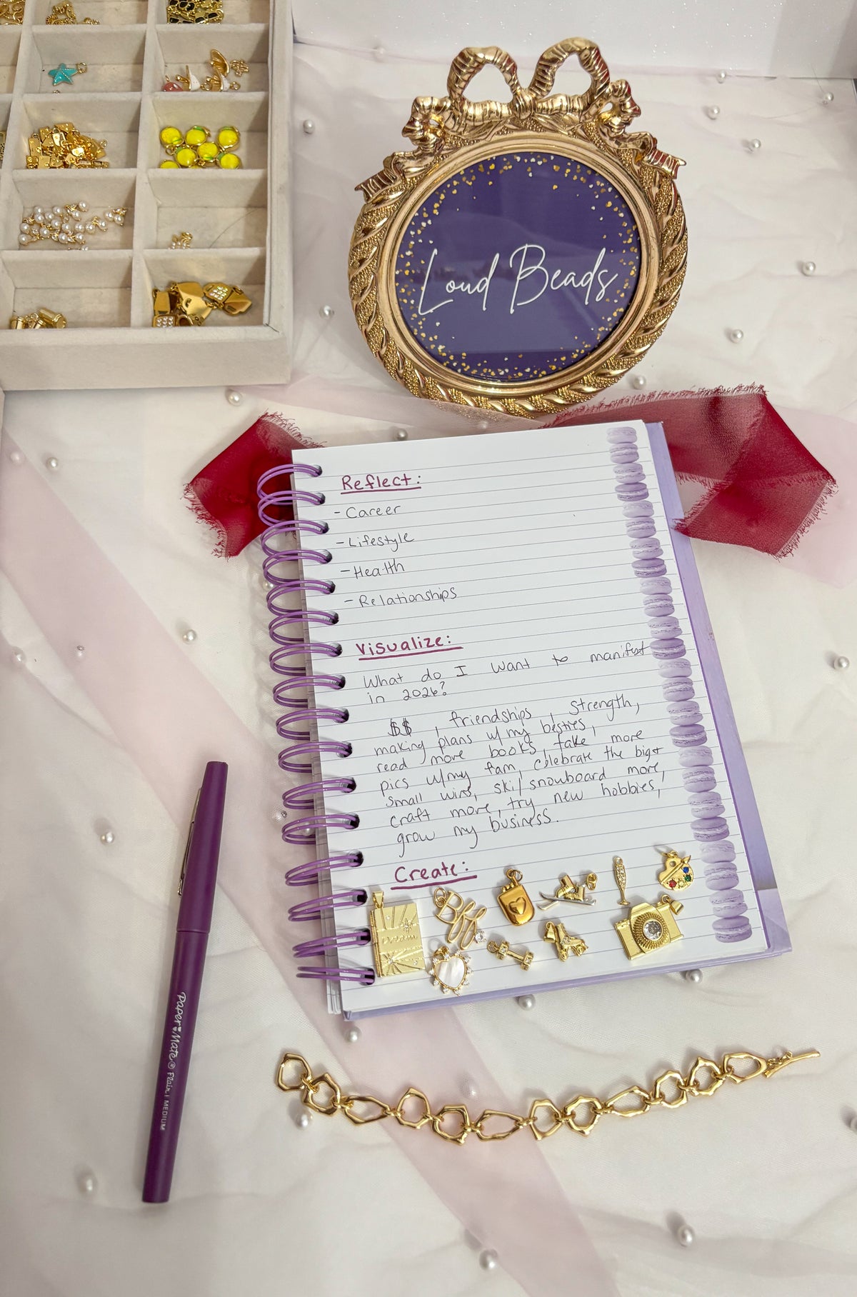 A styled flat-lay featuring a purple spiral-bound journal with handwritten prompts, decorated with gold charms, a purple pen, a gold chain, and a decorative gold-framed mirror on a white fabric background.