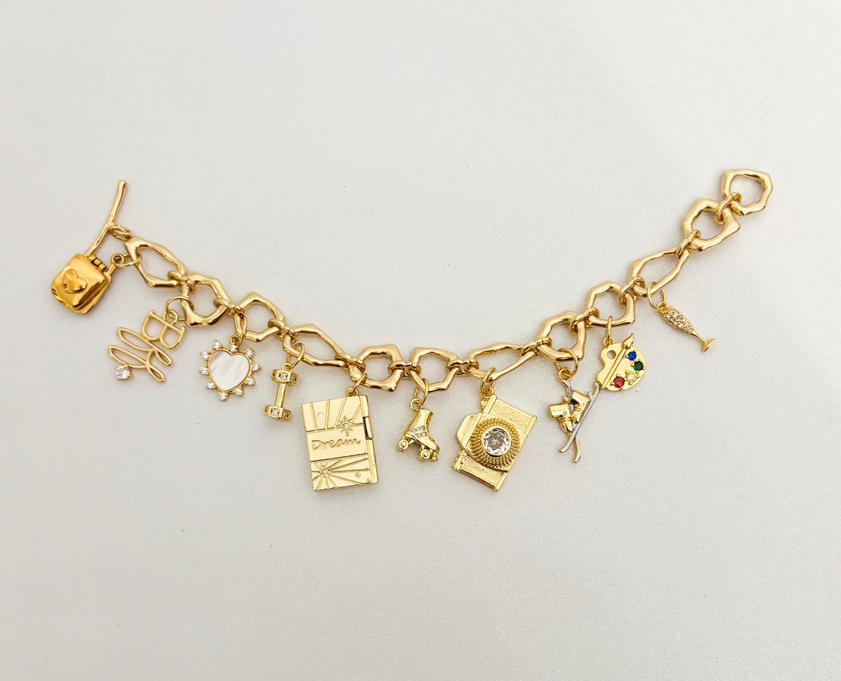 A gold charm bracelet featuring multiple small themed charms, including a film reel, script, camera, paint palette, and other artistic and media symbols, displayed on a light gray background.