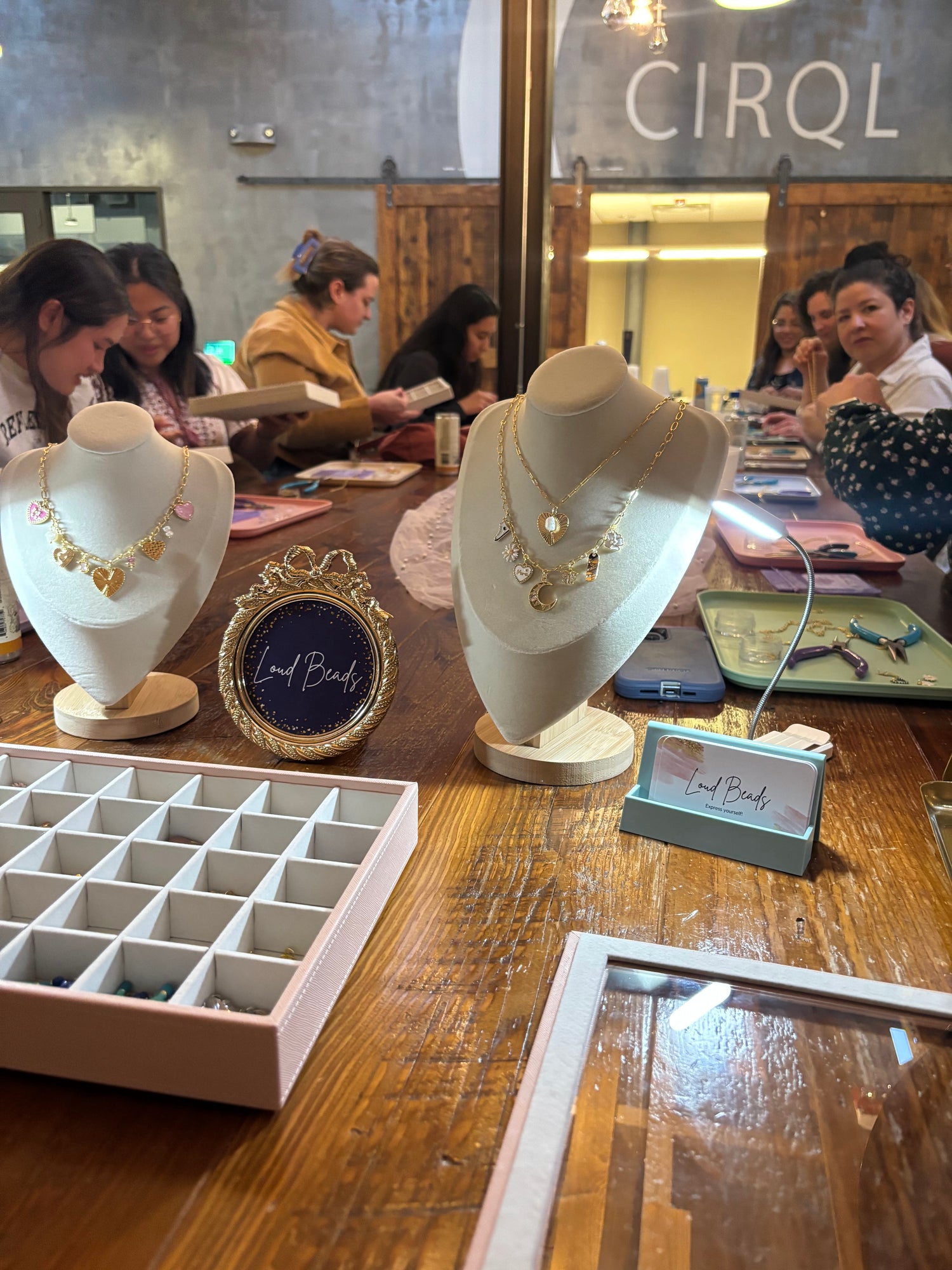 Jewelry display on mannequin heads with women creating jewelry at a table in the background.