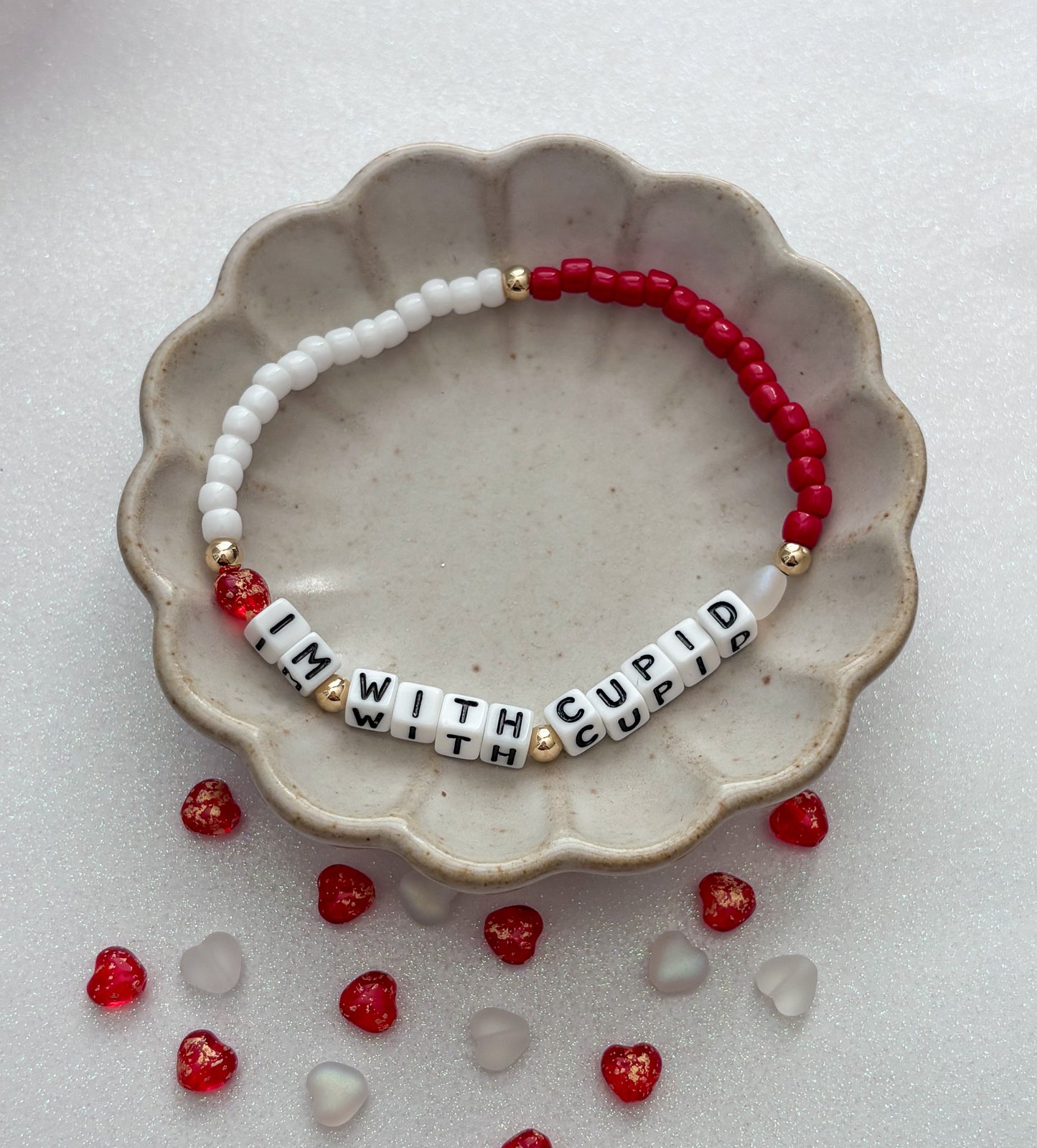 Bracelet with letter beads spelling 'I'm with Cupid' on a ceramic dish with red and clear heart-shaped beads.