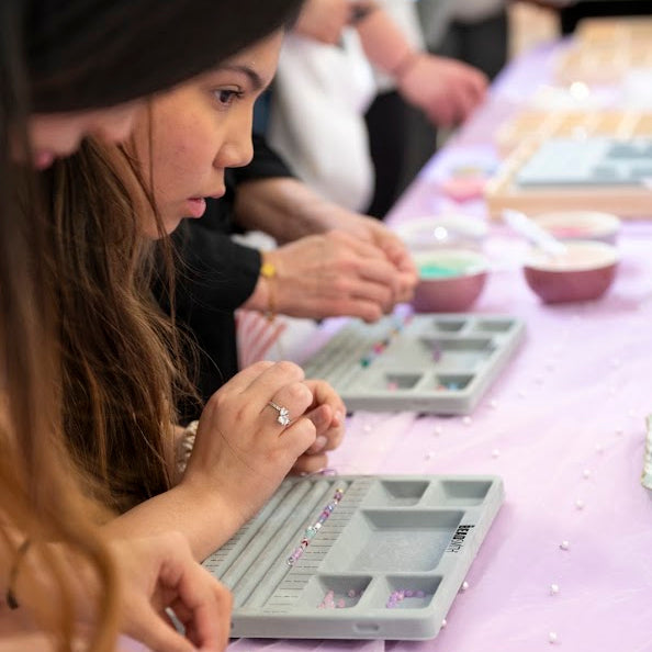 A close-up lifestyle shot of a woman and others working at a craft table, assembling gray plastic jewelry trays with small beads and rhinestones on a light purple tablecloth.