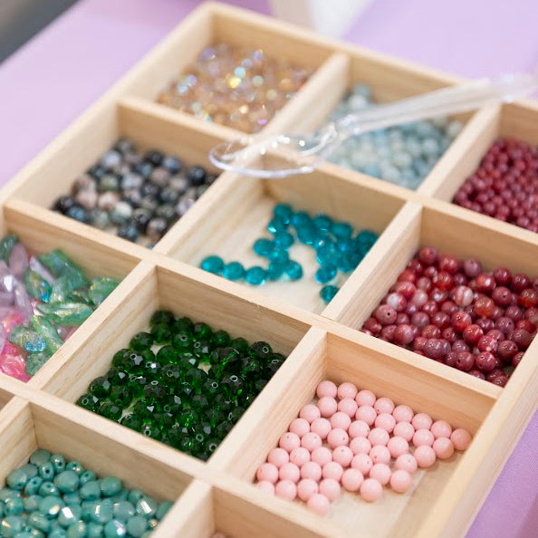 A wooden compartment tray filled with assorted round beads in multiple colors, including pink, teal, green, red, blue, and metallic shades, photographed on a lavender surface.