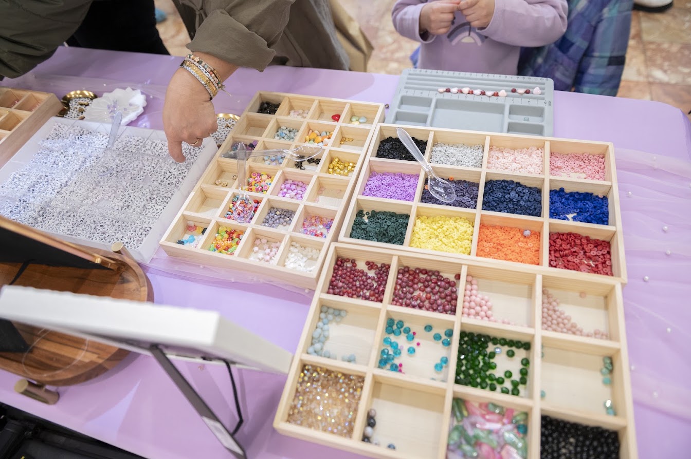 A close-up of a craft table filled with wooden bead trays containing many colorful beads and small embellishments, with a person's hand reaching in and another person standing nearby.